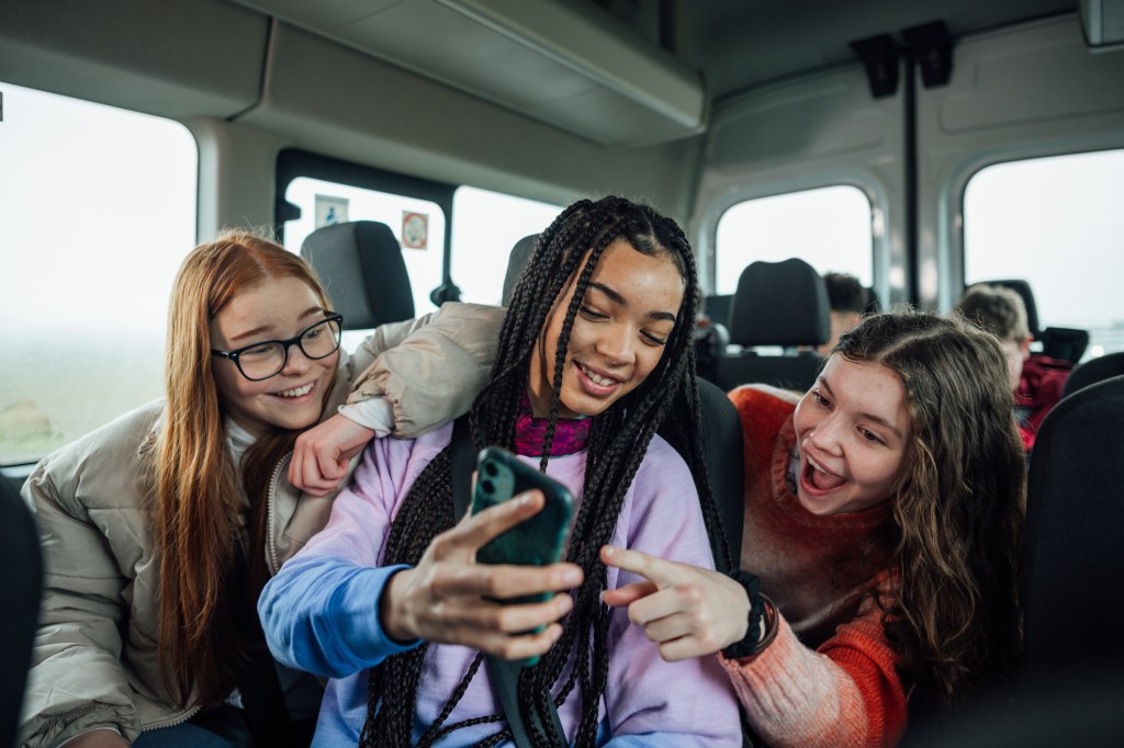 A front view of a group of three teenage girls on the minibus on the way to go on a hiking field trip. They are looking at one of the girls mobile phones and smiling and pointing at the screen.