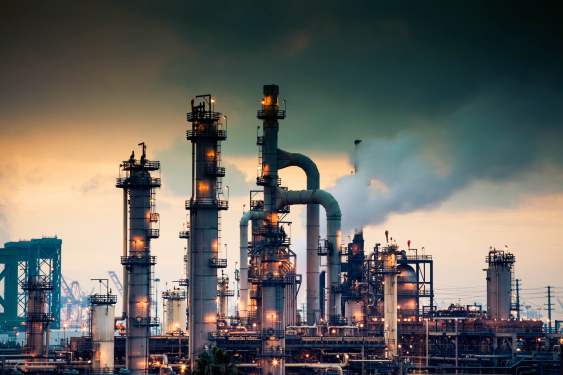 A steaming, glowing mass of smoke stacks and distillation towers connected by conduit and pipe racks towers in the early morning light in front of the Port of Los Angeles.