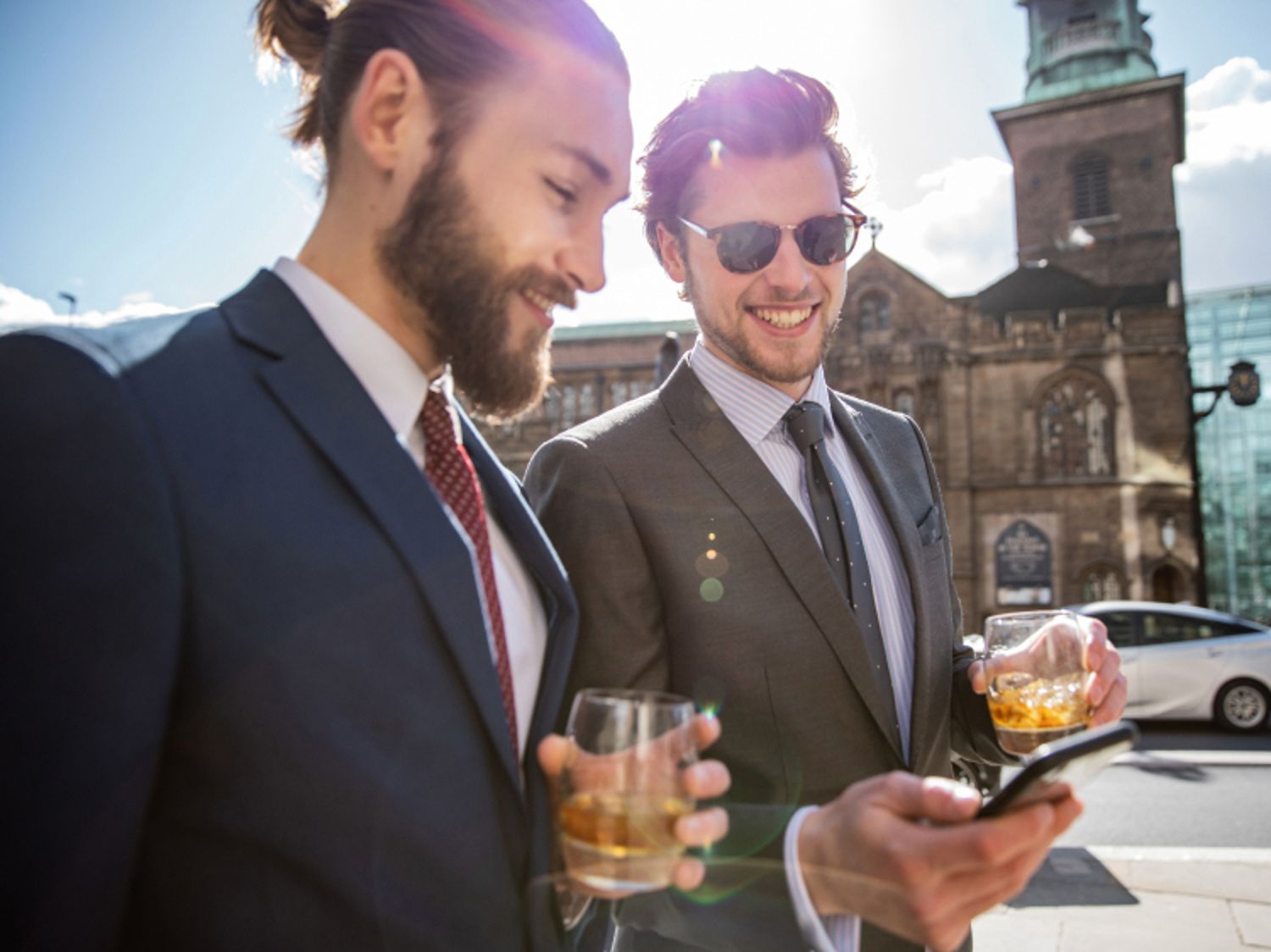 晴れた日の屋外で、飲み物を片手に笑顔でスマホを眺めているスーツ姿の2人の男性