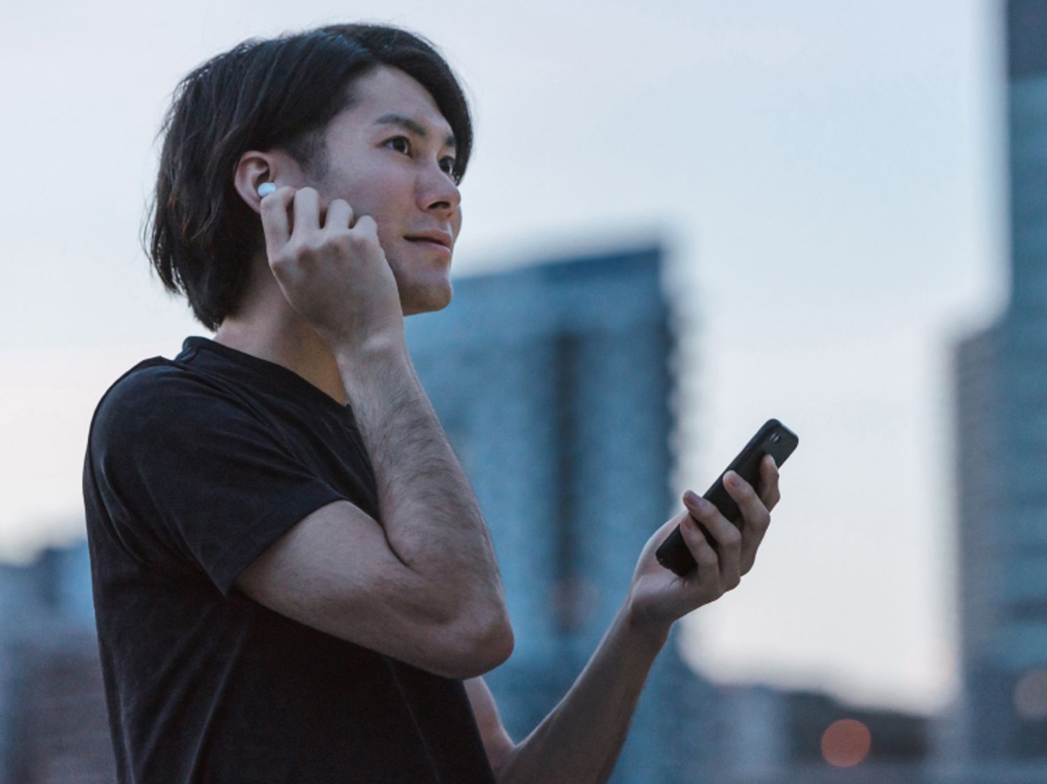 A person outdoors holds a smartphone, wearing a black shirt with a cityscape in the soft evening light.