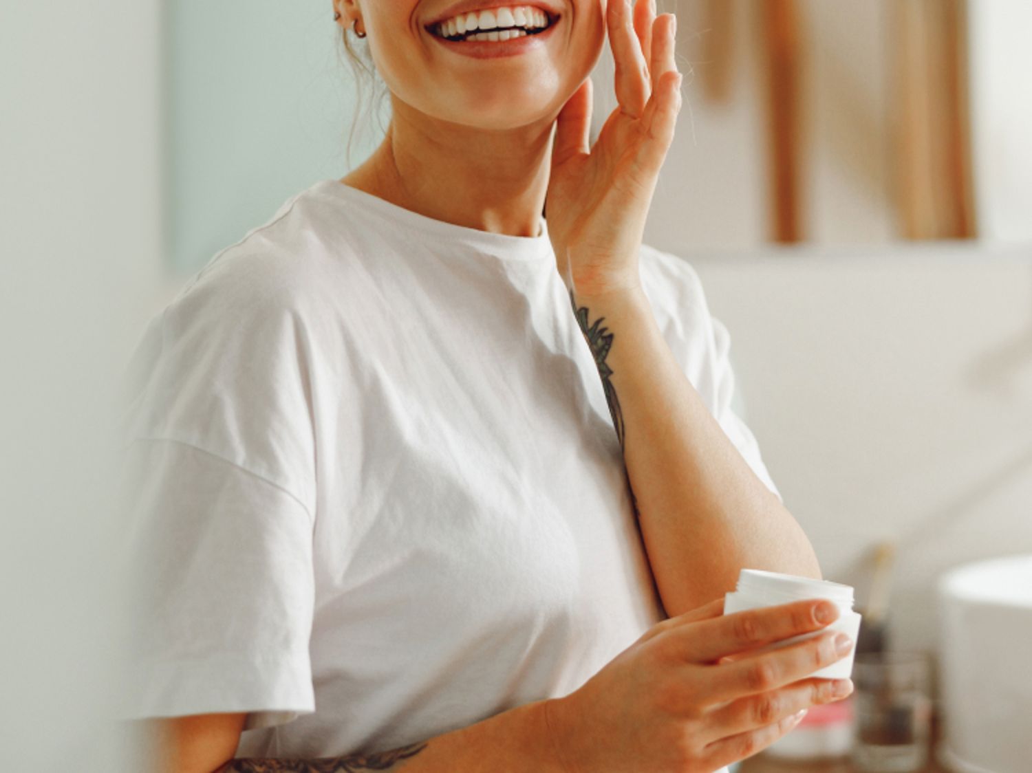 Smiling person applying moisturizer to their face while holding a skincare product.