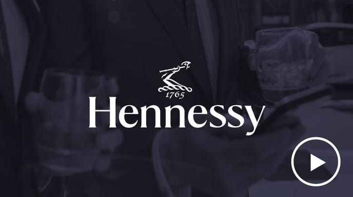 Two men in suits holding glasses of Hennessy while looking at a smartphone, overlaid with the Hennessy logo and a play button.