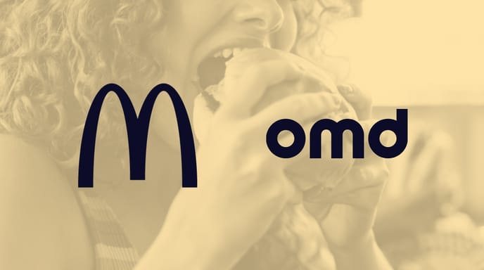 Close-up of a person with curly red hair taking a bite of a sandwich, smiling in a bright, casual setting.