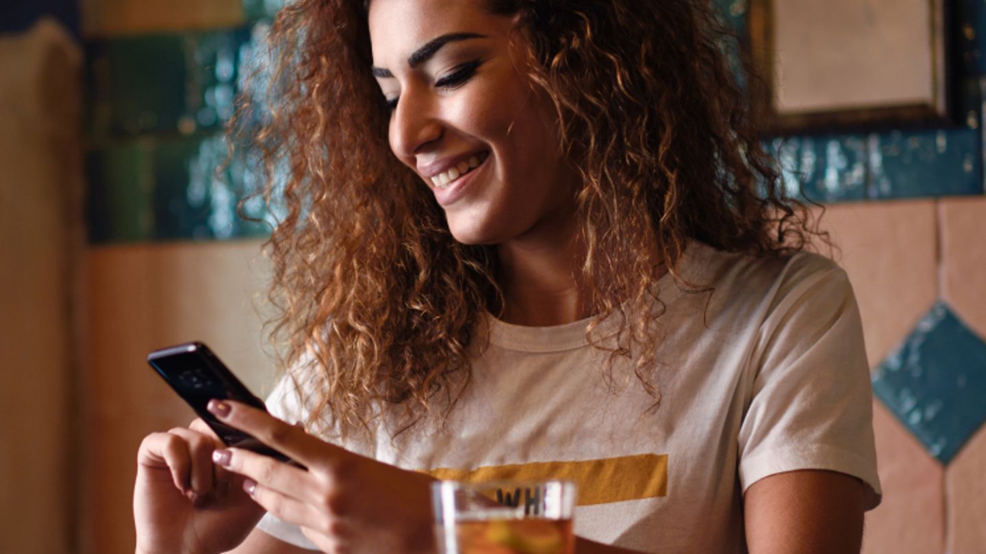 A smiling woman with curly hair sits at a café table, looking at her phone with a glass of iced tea in front of her
