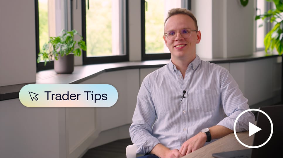 In einem hellen, modernen Büro mit Pflanzen und großen Fenstern sitzt eine lächelnde Person mit Brille und hellblauem Hemd. Auf dem Bildschirm sind eine Grafik mit der Aufschrift „Trader Tips“ und ein Play-Button-Symbol zu sehen.
