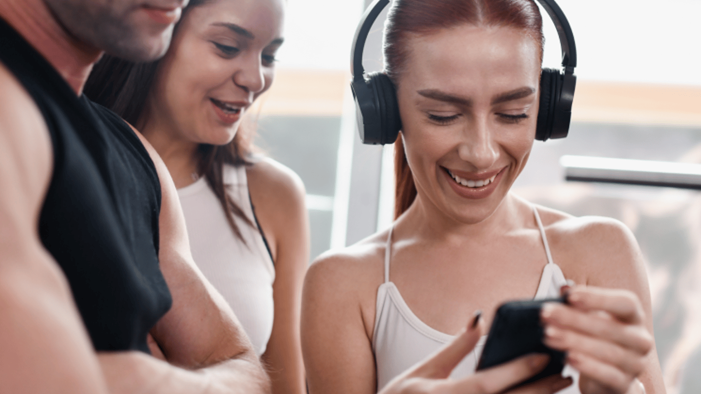 A group of three fit individuals at the gym, with a smiling woman wearing headphones showing something on her phone to the others.
