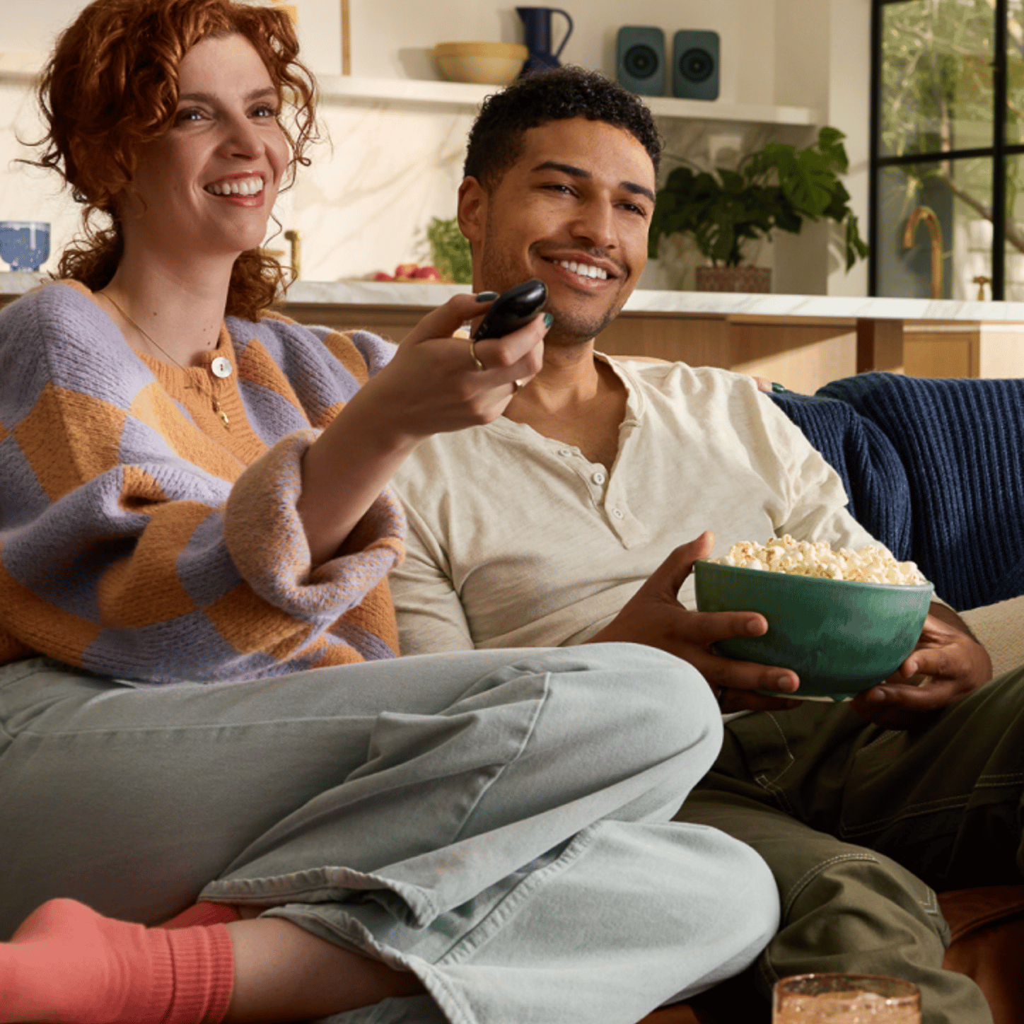 Una alegre pareja está sentada en el sofá con el mando a distancia y un cubo de palomitas, disfrutando de un momento de relax mientras ven juntos la tele en una moderna y acogedora sala de estar.