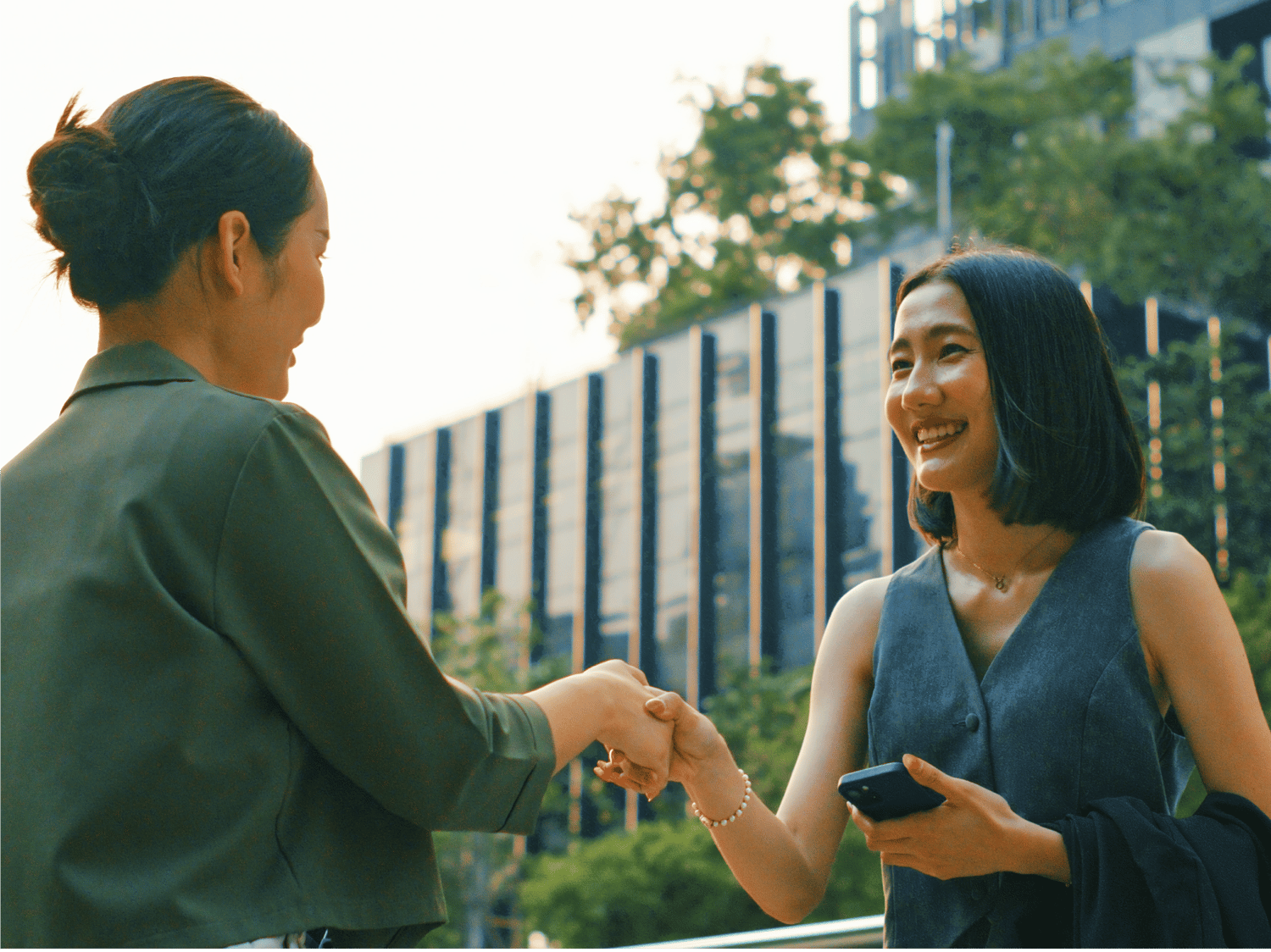 Two women smile and shake hands outdoors in a modern urban setting, one holding a phone and wearing a sleeveless outfit, with office buildings and greenery in the background.