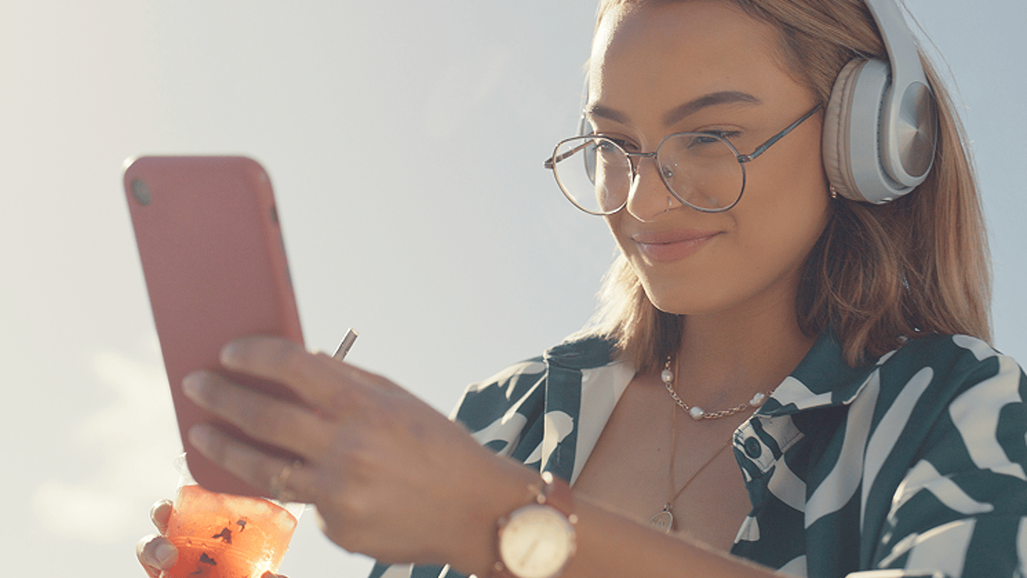 Woman wearing headphones using smartphone outdoors while enjoying a drink